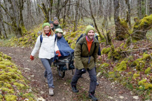 photo couleur d'une randonnée solidaire en Périgord, montrant un handicapé ou PMR sur une Joëlette tractée par des bénévoles de l'association ÇA ROULE POUR TOI sur un chemin forestier de Dordogne